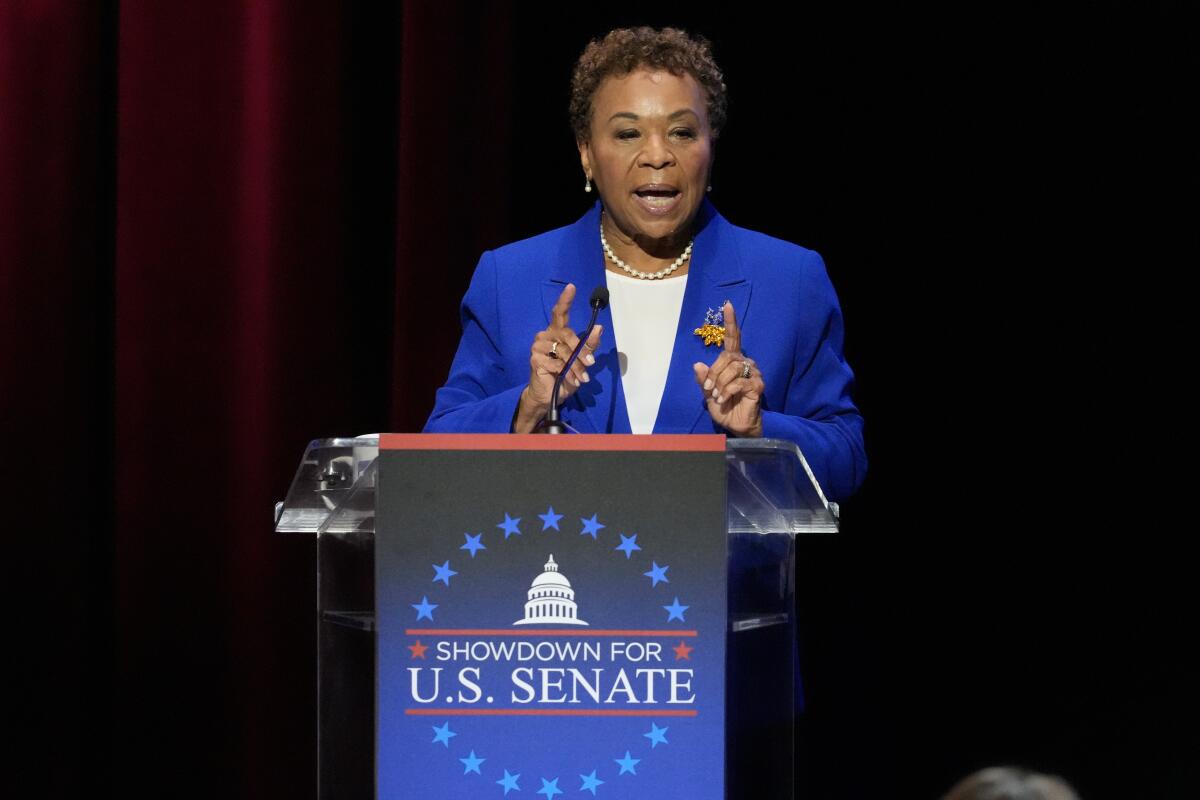 Barbara Lee, in a blue blazer, gestures while speaking from a lectern, its blue sign reading, "Showdown for U.S. Senate"