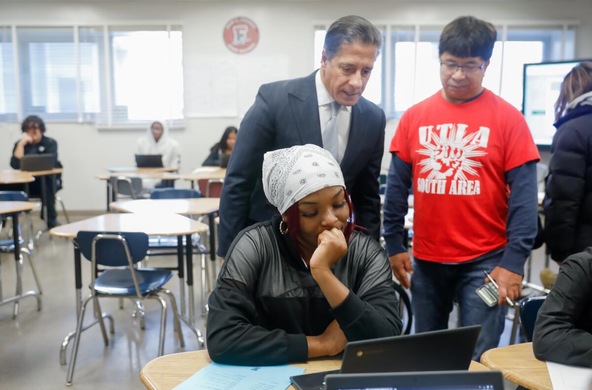 Two men, one in a suit and the other in a UTLA shirt, watch a student working on a laptop.