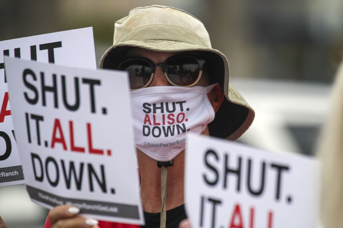 A protester in a "Shut Aliso Down" mask holds signs reading "Shut It All Down."