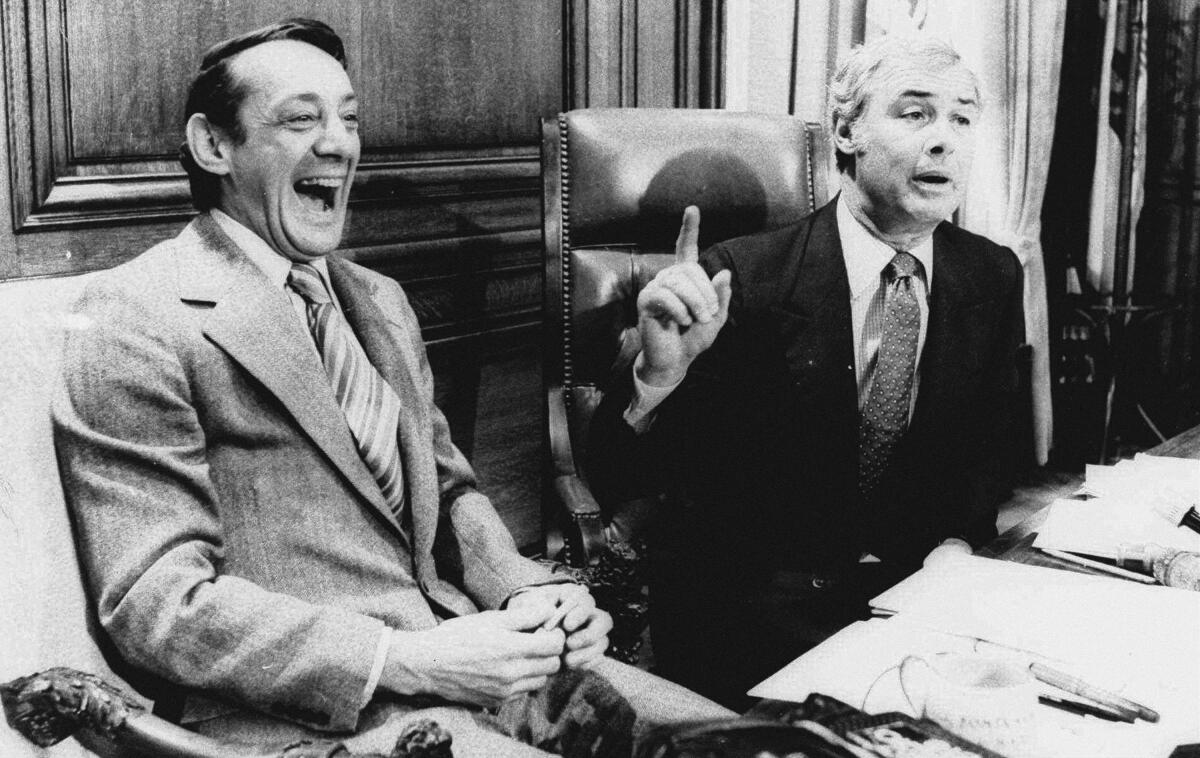 A black-and-white photo of two men sitting at a table with paper piles, one laughing as the other raises a finger and speaks.