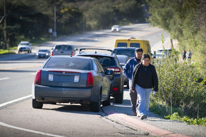 RAMONA, CA - JANUARY 26: Pedro Avendano, and Arcelia Dominguez, foreground, from Oceanside, walk next to parked vehicles on the shoulder of Highway 67 near the Mount Woodson Trail, as they head for the trial and a hike up to Potato Chip Rock Thursday January 26, 2023. The San Diego County Board of Supervisors has decided to move forward with a plan to construct a parking lot for the hikers. (Howard Lipin / For The San Diego Union-Tribune)