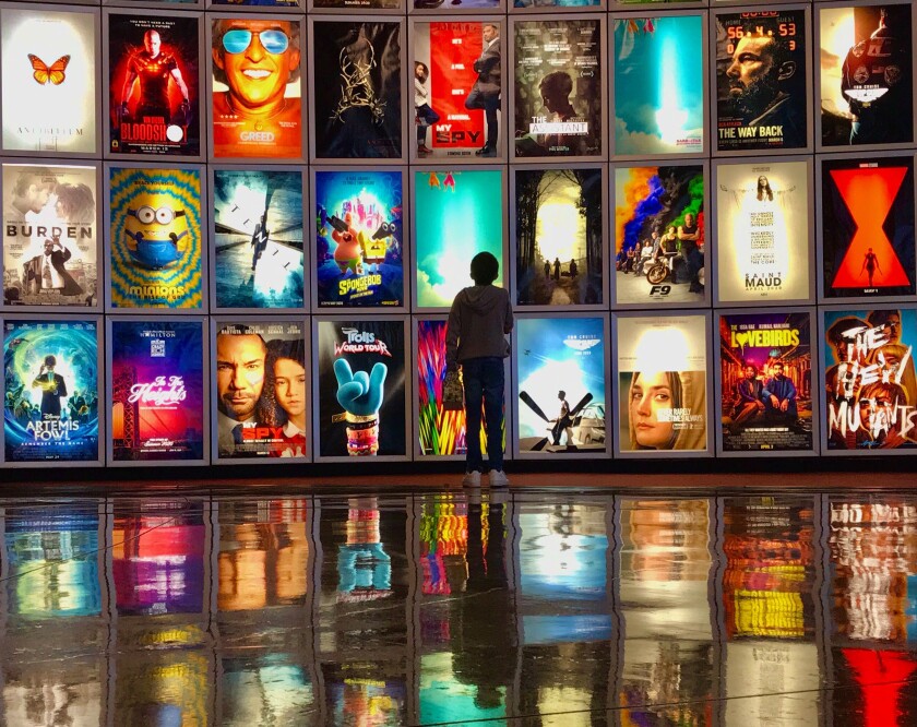 A child stands in front of a wall of movie posters reflected on a polished floor.