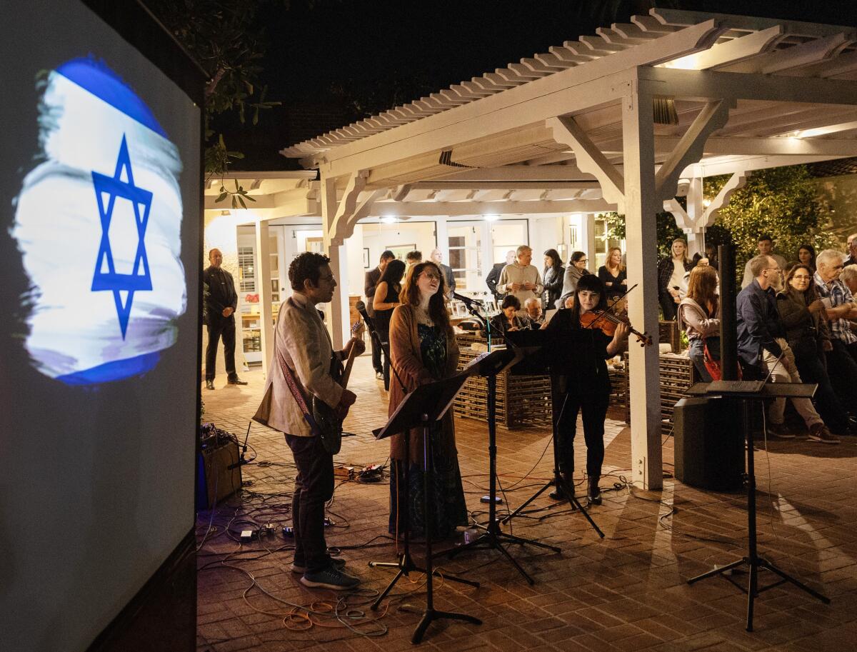 A group performs music near an image on a screen of the Israeli flag while people gather in the background.