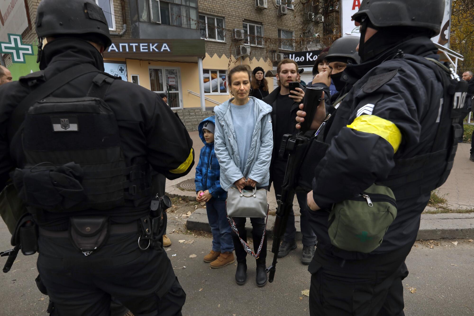 A woman in a light blue jacket and top and holding a purse is surrounded by men in helmets and dark uniforms on a street