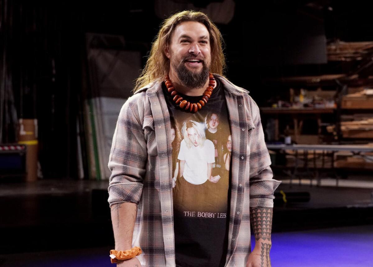 Jason Momoa in a flannel shirt and black T-shirt standing on a stage smiling.