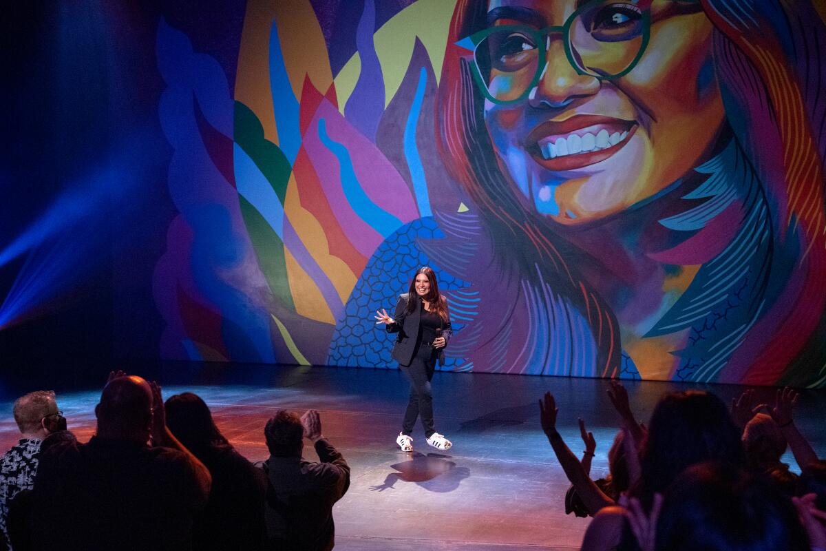 Cristela Alonzo doing a stand-up comedy performance in front of a mural.