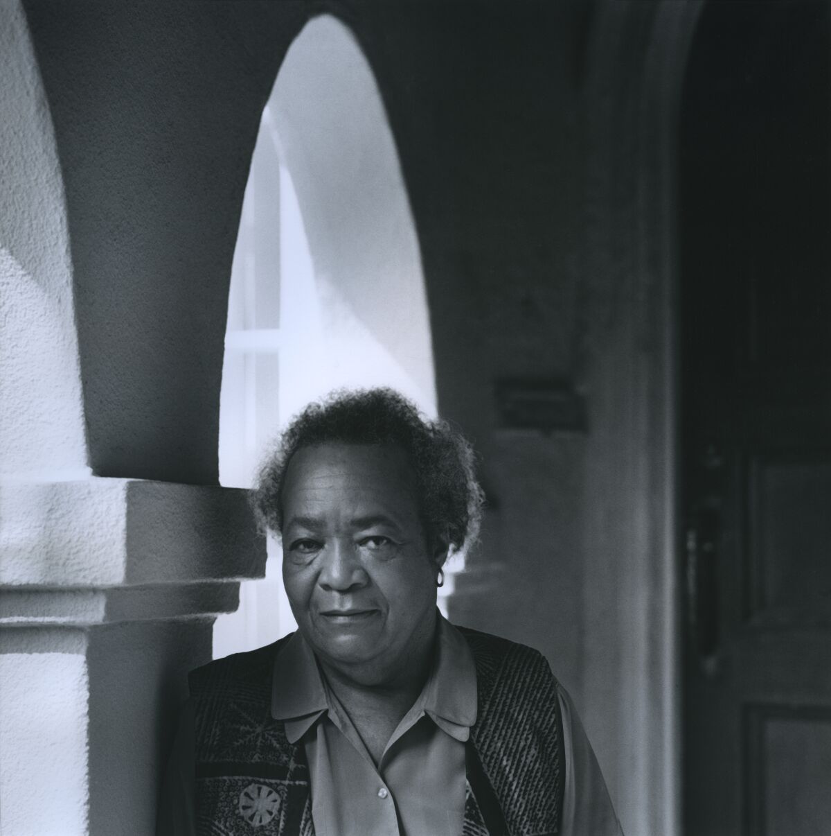 Samella Lewis is seen in a black and white photo leaning against a column and framed by an arch