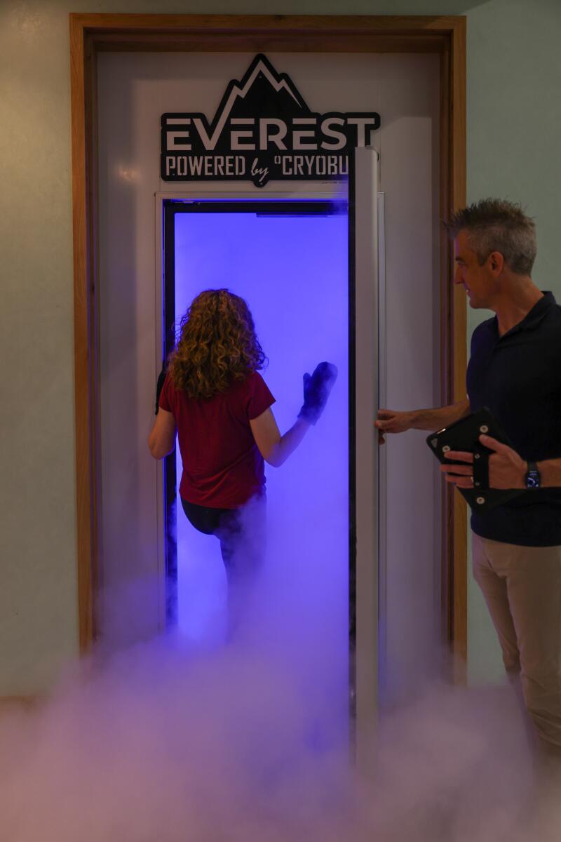 Cold vapor billows out of a cryotherapy chamber as a woman steps into it. A man stands by the open door.