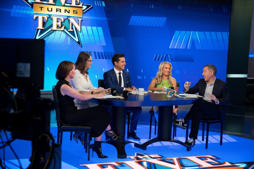 Three women and two men seated at a table on a TV show set.