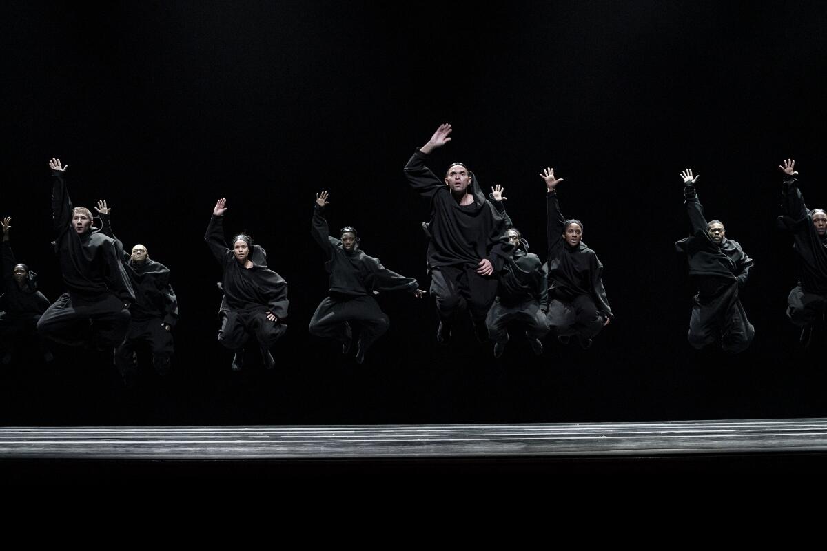 A group of dancers, dressed in charcoal gray ensembles, jump on stage in perfect formation.