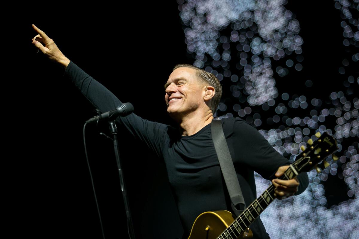 A smiling Bryan Adams points to the sky and holds his guitar while performing onstage