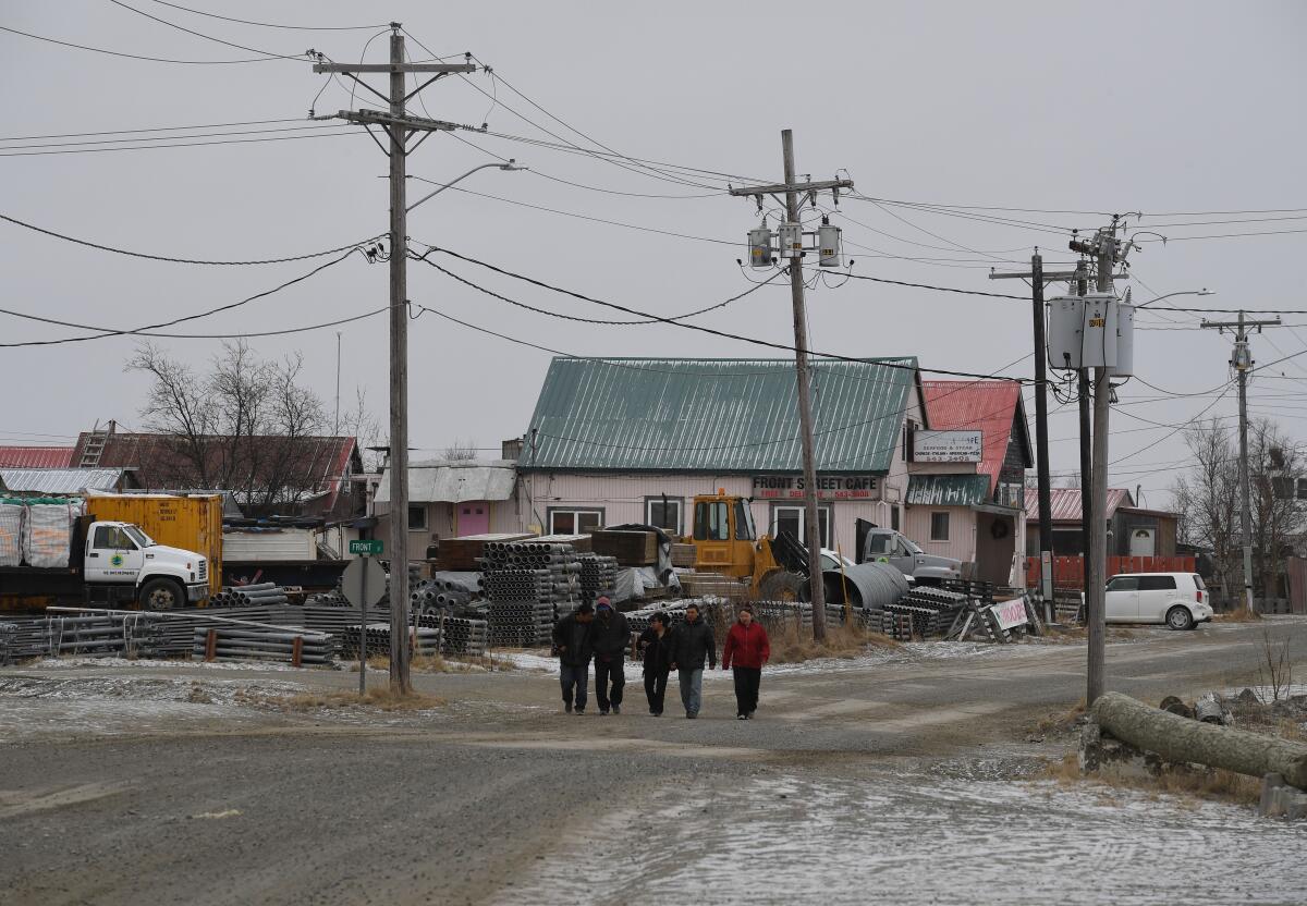 A small group of people walks past weather-worn buildings and an industrial yard on a dirt road.