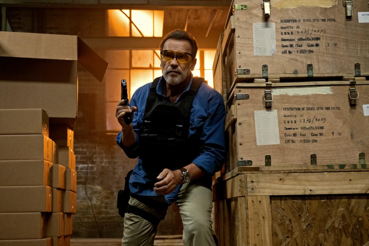 Arnold Schwarzenegger hides behind crates wearing a bulletproof vest and holding a gun