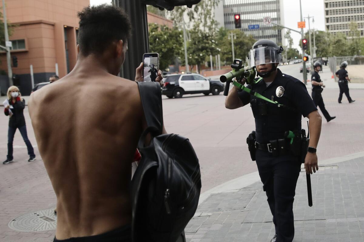 An officer in dark uniform and face shield holds a launcher on his shoulder aimed at a shirtless man holding up a cellphone 