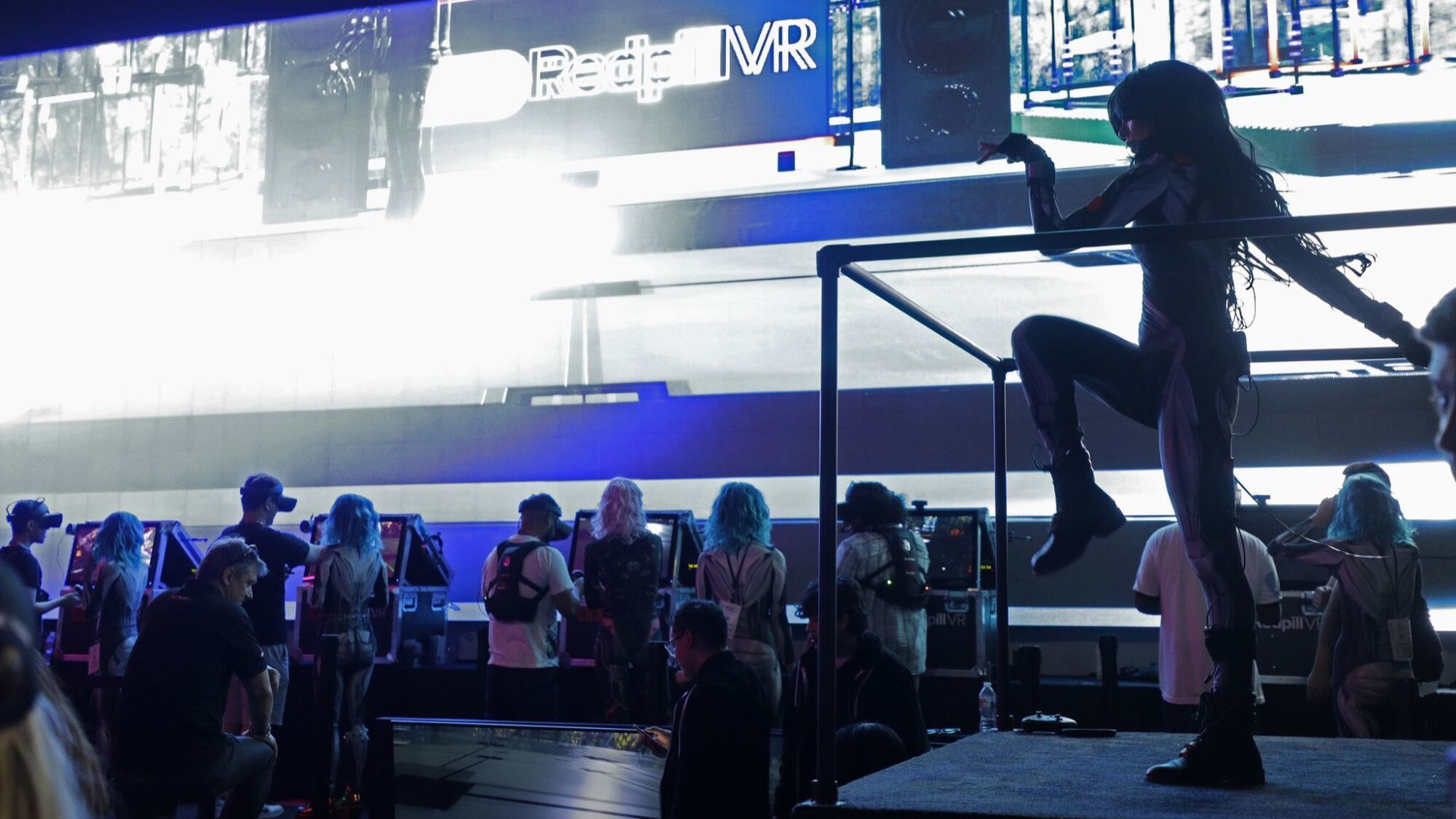 A woman dances on a platform as people gather at consoles below a bank of video monitors