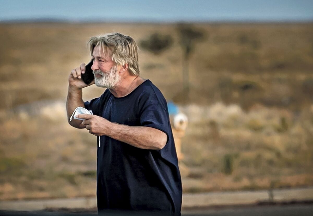 Alec Baldwin outdoors talking on a cellphone and holding a face mask.