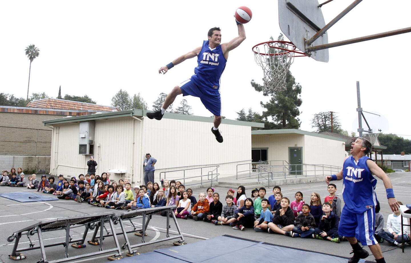 Keith Cousino of the TNT Dunk Squad goes up in the sky for a dunk as partner Brian Smith, right, looks on along with La Canada Elementary School kindergarten through 3rd grade students at the school playground in La Canada Flintridge on Tuesday, March 25, 2014.