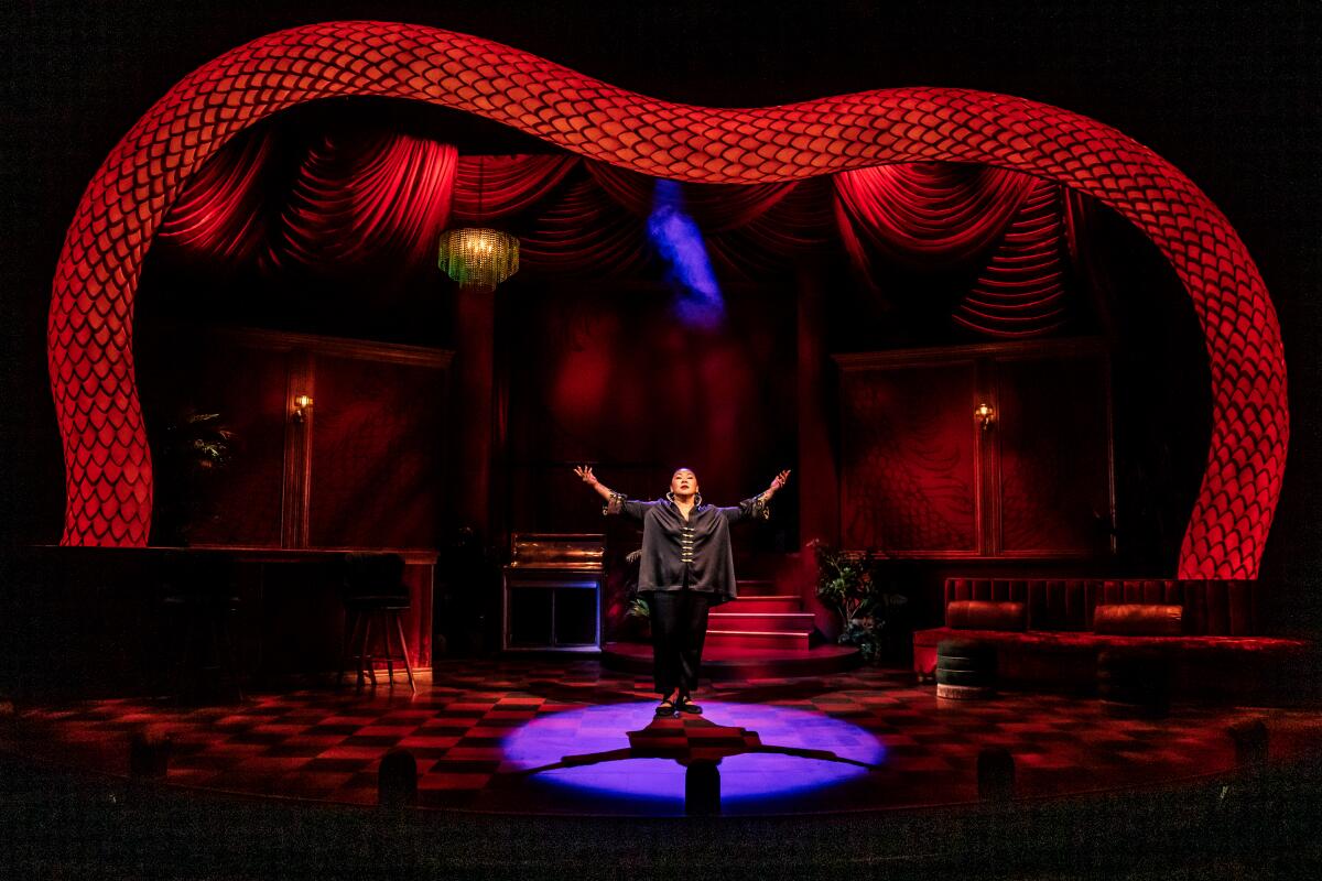 A woman stands, arms upraised, on a red-lighted stage under a frame invoking a red snake