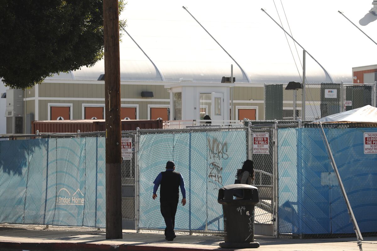 A person walks along a city sidewalk past a chain-link fence lined with blue plastic sheeting