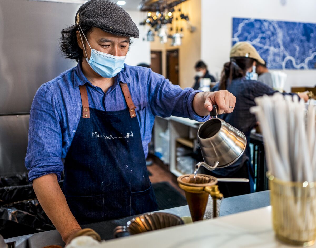 A man wears an apron and makes coffee at a coffee shop.