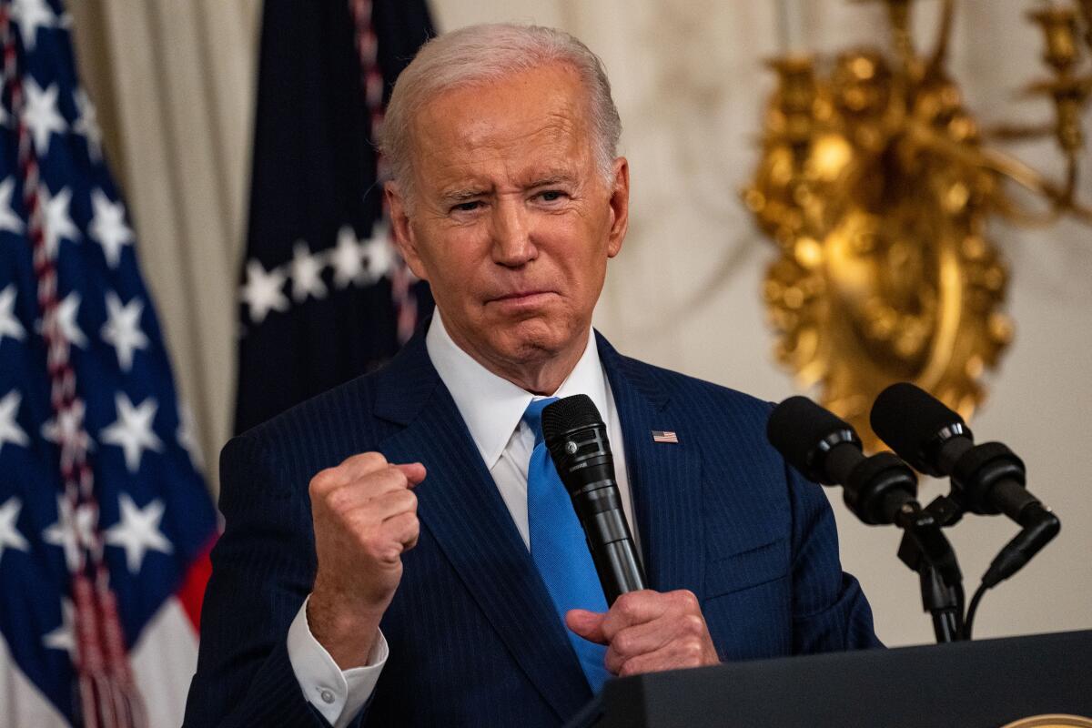 President Biden clenching his right hand and holding a microphone in his left as he stands at a lectern in an ornate room.  