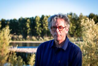 An older man in red rimmed glasses standing in front of a lake lined with evergreens.