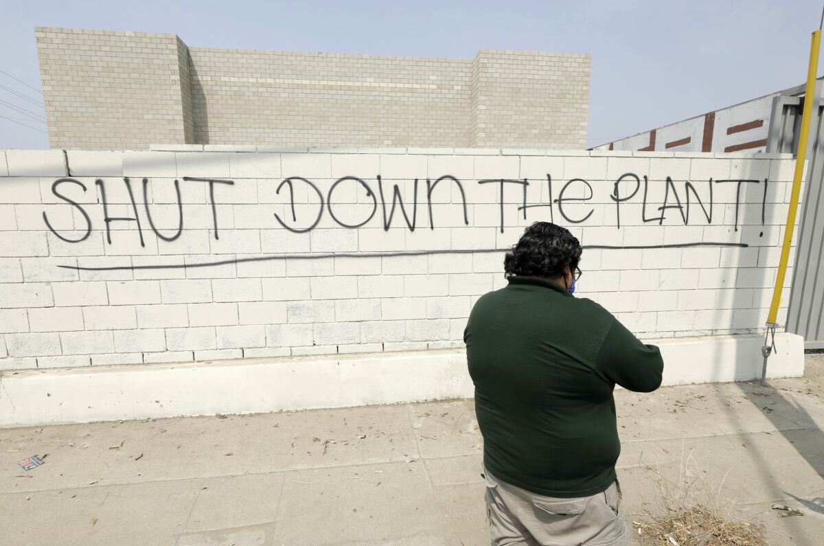 A man stands in front of a cinder-block wall on which someone has scrawled "SHUT DOWN THE PLANT!"