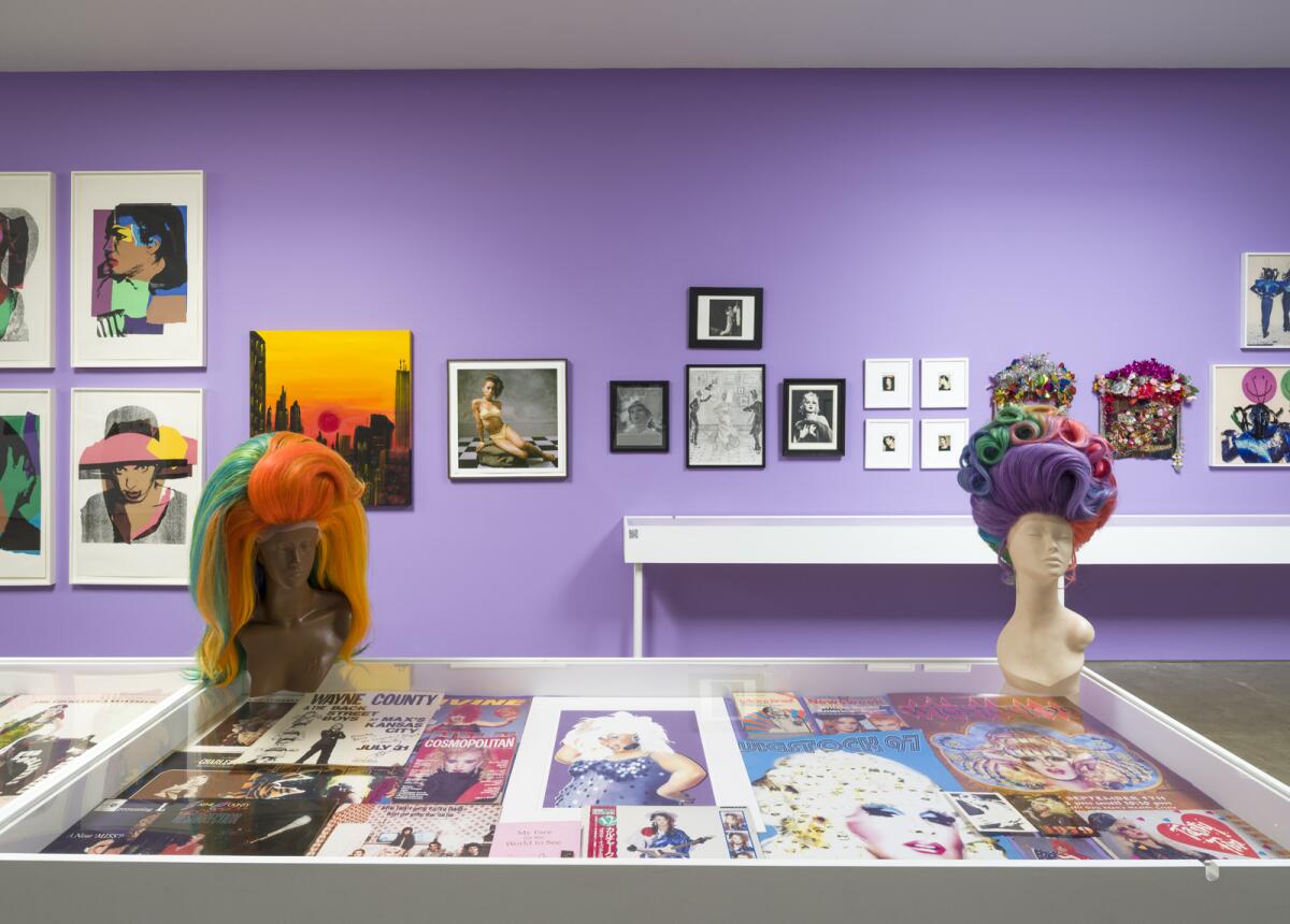 A gallery with lavender walls contains vitrines with drag ephemera and two mannequins wearing flamboyant rainbow wigs