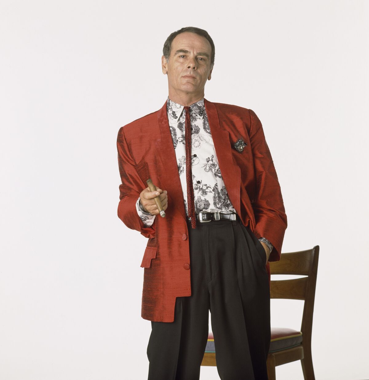 A man in a geometric red blazer, black trousers, patterned shirt and skinny tie holding a cigar.