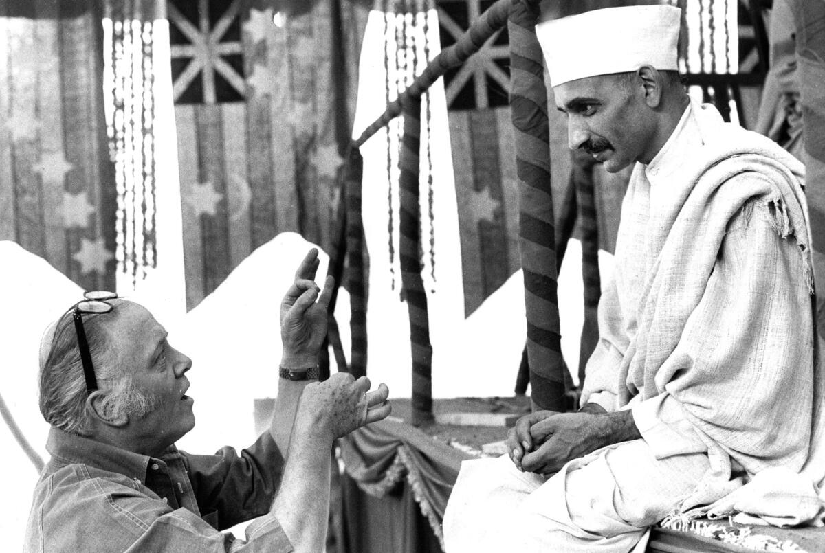 A man with glasses up on his forehead kneels and gestures before a seated actor dressed as Gandhi