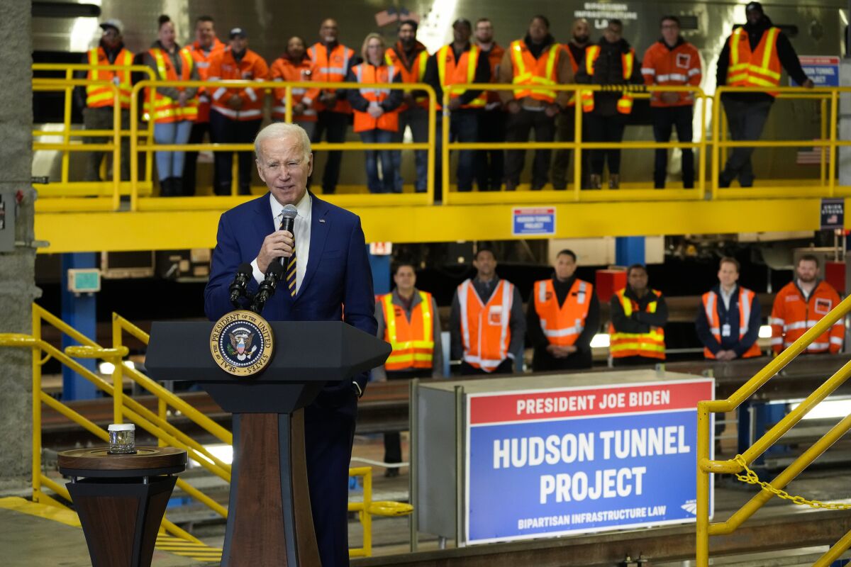 A white-haired man in a blue suit speaks at a lectern. People in construction safety vests are shown in the background.
