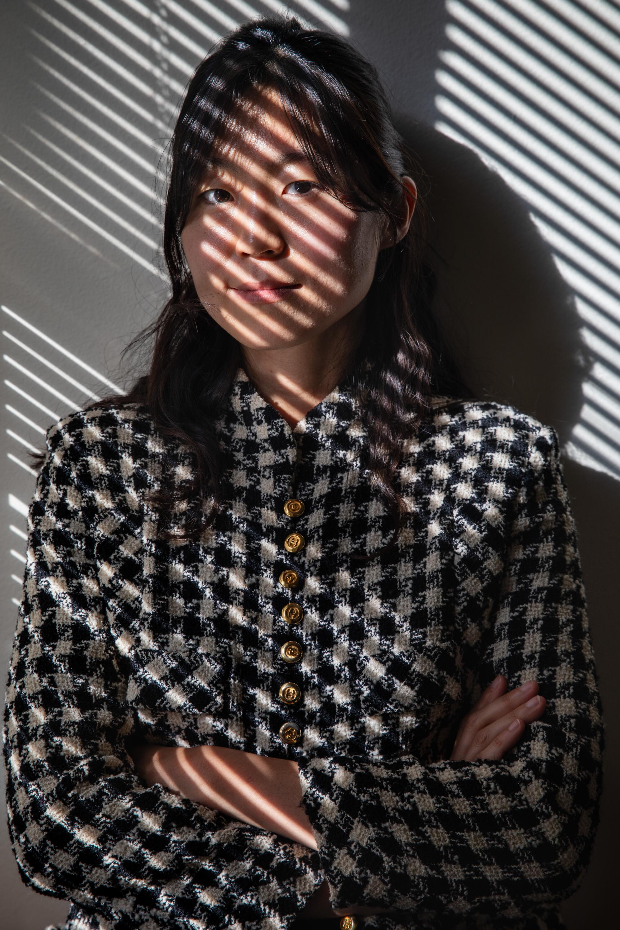 Juyoung Yoon, wearing a houndstooth jacket, sits near a window, lighted by a striped light streaming in through blinds