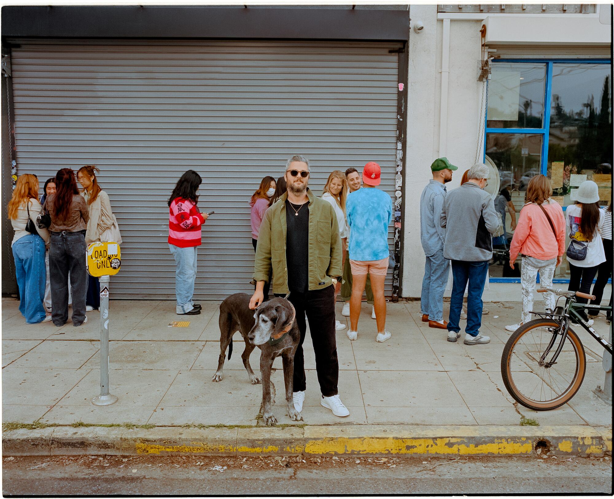Man in a green jacket stands with his dog on the sidewalk.