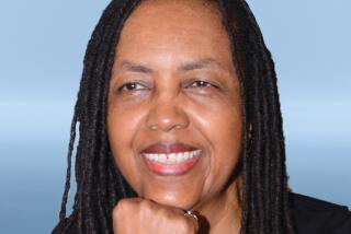 A woman with dreadlocks in a black long-sleeved shirt sits in front of a generic blue background for a portrait.
