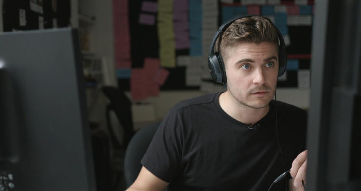 A man in a black T shirt and head phones sits in front of a computer