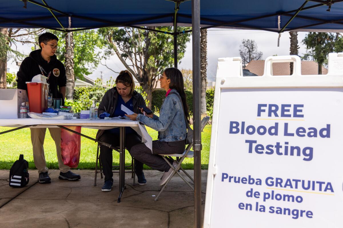Three women sit at a table under a tent. A sign says "Free blood lead testing."