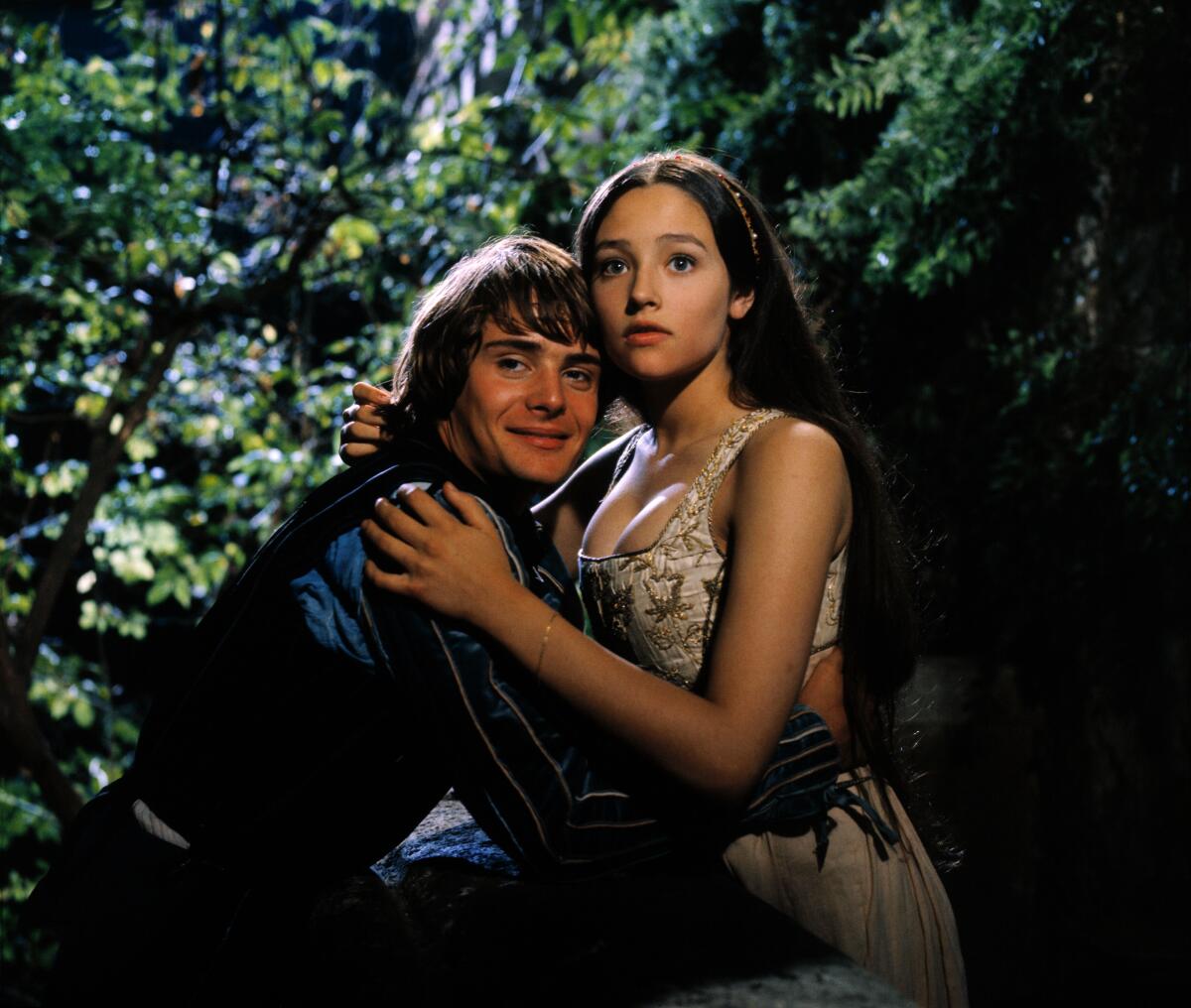 A boy and a girl in old-fashioned formal attire embrace in a forest.