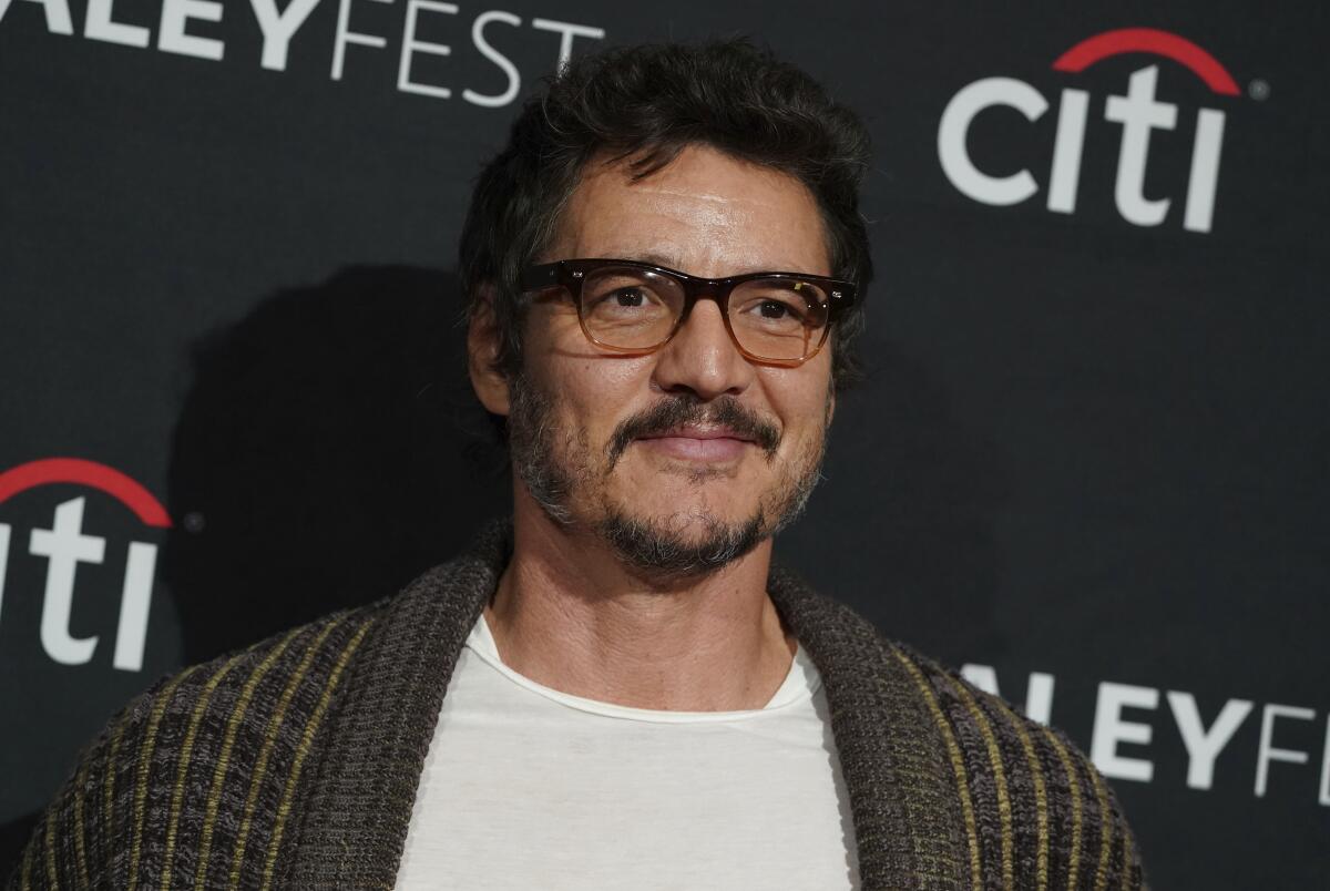 Pedro Pascal wears a white undershirt with a green knit sweater while posing for a photo at a red carpet event. 