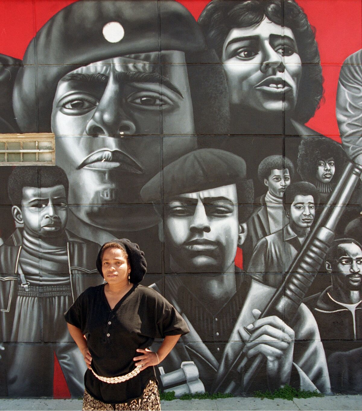 Artist Noni Olabisi stands before a mural of Black Panthers set against a red backdrop 