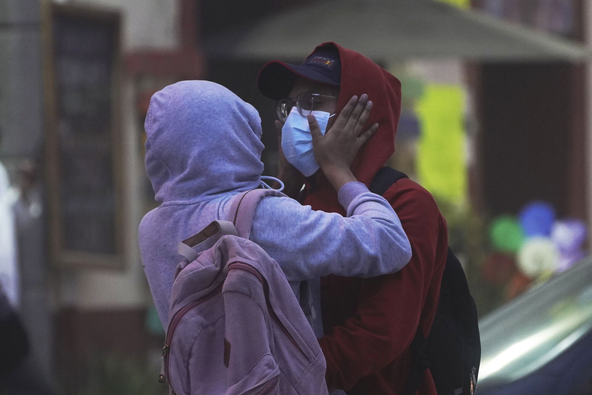 A person in a lavender hoodie and pink backpack places hands on the masked face of a person in a red hoodie, wearing glasses 
