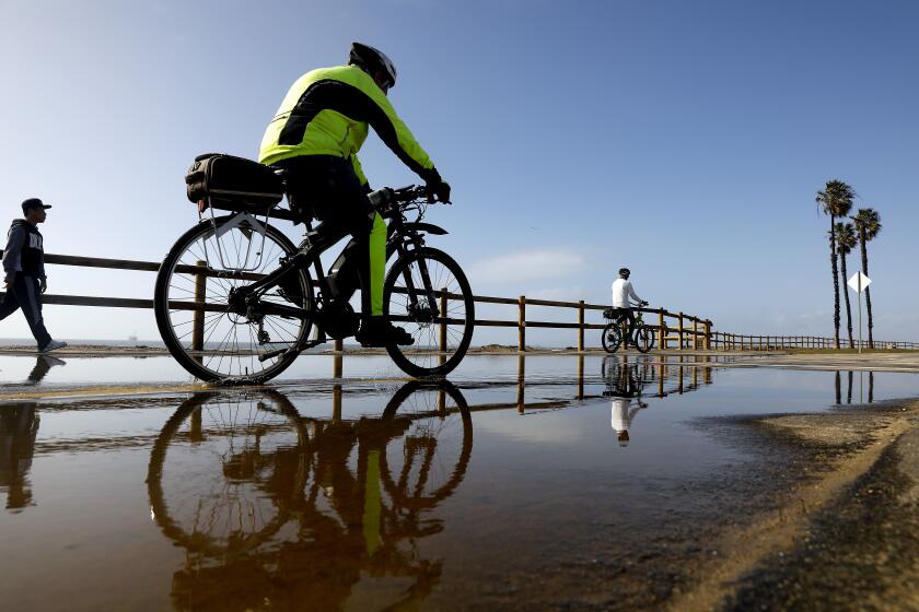 HUNTINGTON BEACH-CA-JANUARY 5, 2023: Cyclists and pedestrians are reflected in puddles along a pathway off Pacific Coast Highway in Huntington Beach on Thursday, January 5, 2023 near where PCH between Warner Avenue and Seapoint Drive is closed due to flooding. (Christina House / Los Angeles Times)