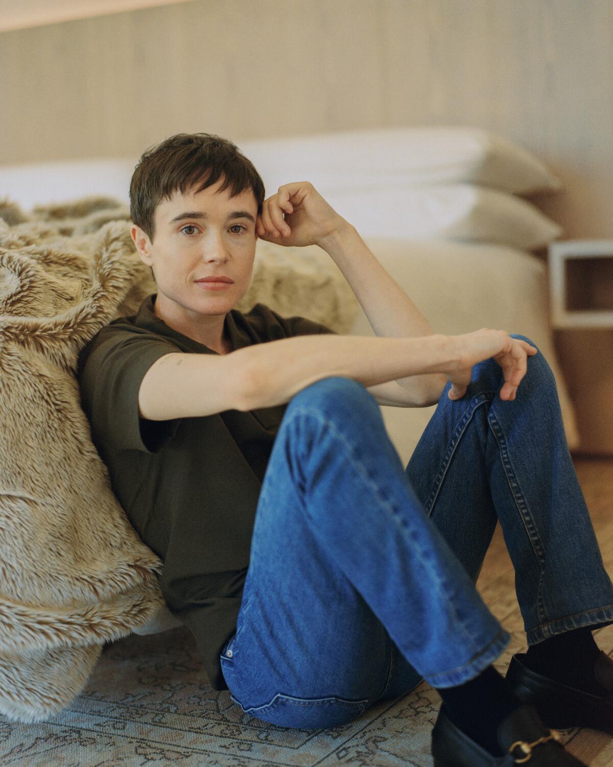Elliot Page sits, leaning against a bed in an olive polo and blue denim jeans.