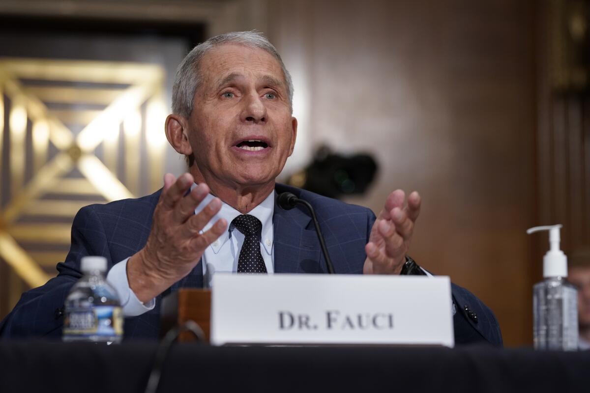 Anthony Fauci, seated at a table behind a nameplate that says "Dr. Fauci," gestures with his hands while speaking.