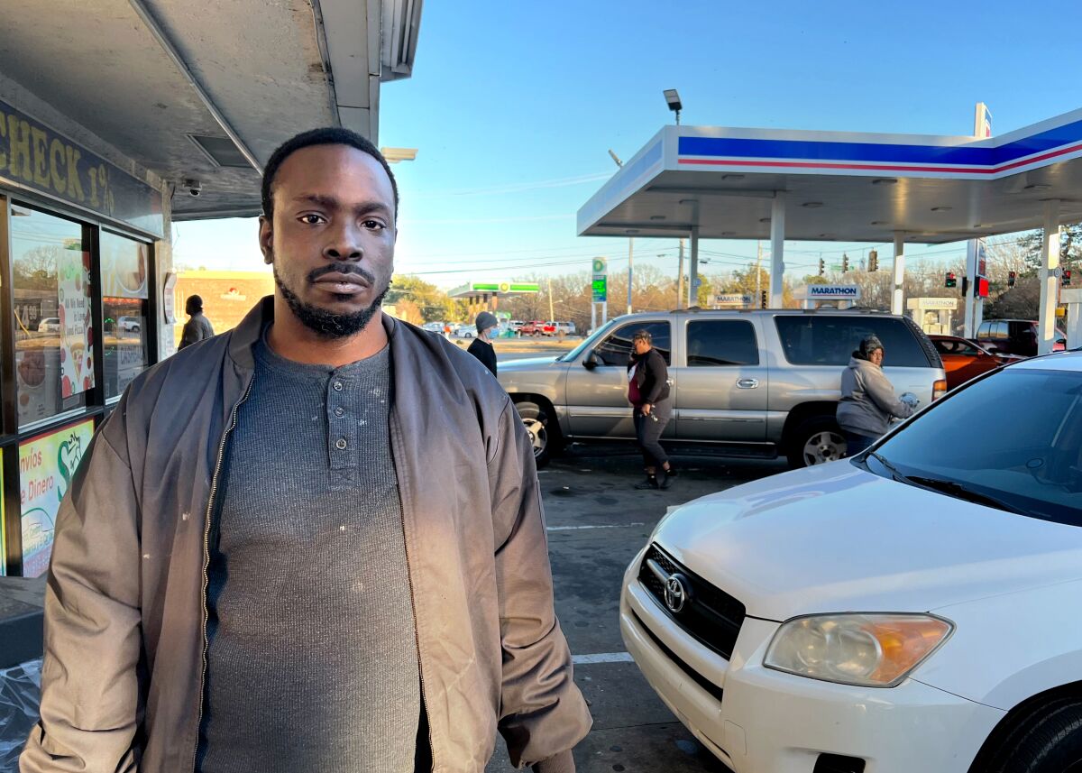 At a gas station, a black man in a brown jacket stands to the left and looks directly at the camera.