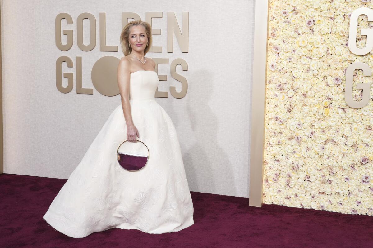 Gillian Anderson in a long white gown holding a burgundy purse posing on a red carpet in front of a Golden Globes sign