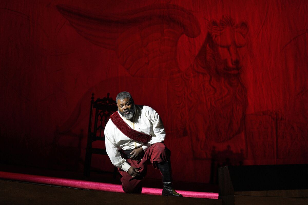 Russell Thomas, wearing red pants and a white shirt, kneels as he sings on a red stage set.