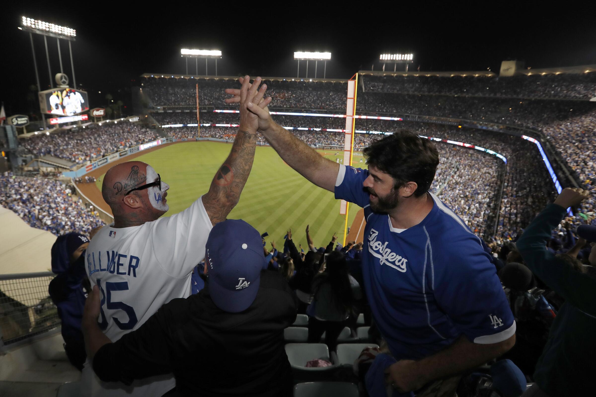 Two fans in Dodgers jerseys, one with Dodgers face paint, high five as fans at Dodger Stadium celebrate after a win