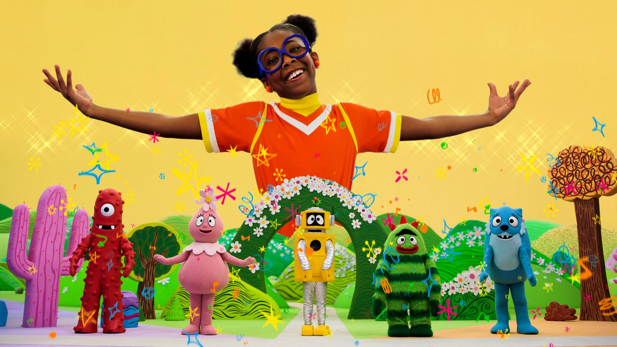 A girl in blue glasses and an orange top stands with her arms extended over five puppets.