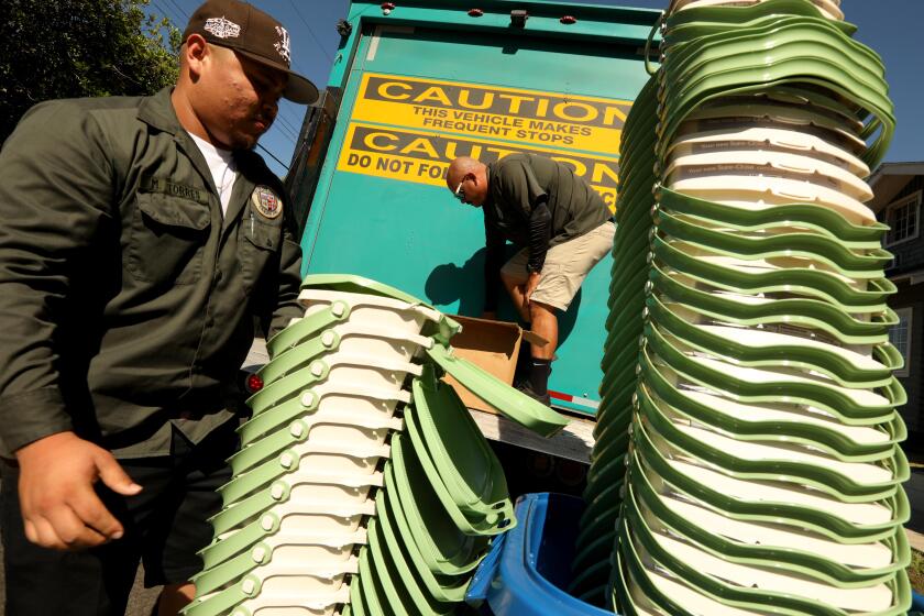 LOS ANGELES, CA - AUGUST 6, 2022 - - LA Sanitation workers Miguel Torres, left, and Zac Ortiz, unload two stacks of in-home composting pails to be handed out to homeowners in Los Feliz on August 6, 2022. 2022 is the year that most residents of Los Angeles are being instructed to dump their kitchen waste into their green organic waste bin. Previously leftover food went into the regular/landfill (brown) trash can. This shift represents the biggest change in solid waste management in decades. The new program comes under a state law that requires cities and counties to change the way they handle leftover food and kitchen waste. The change is designed to help improve soil health and to reduce methane and other greenhouse gases that form when organic waste goes to a regular landfill. (Genaro Molina / Los Angeles Times)