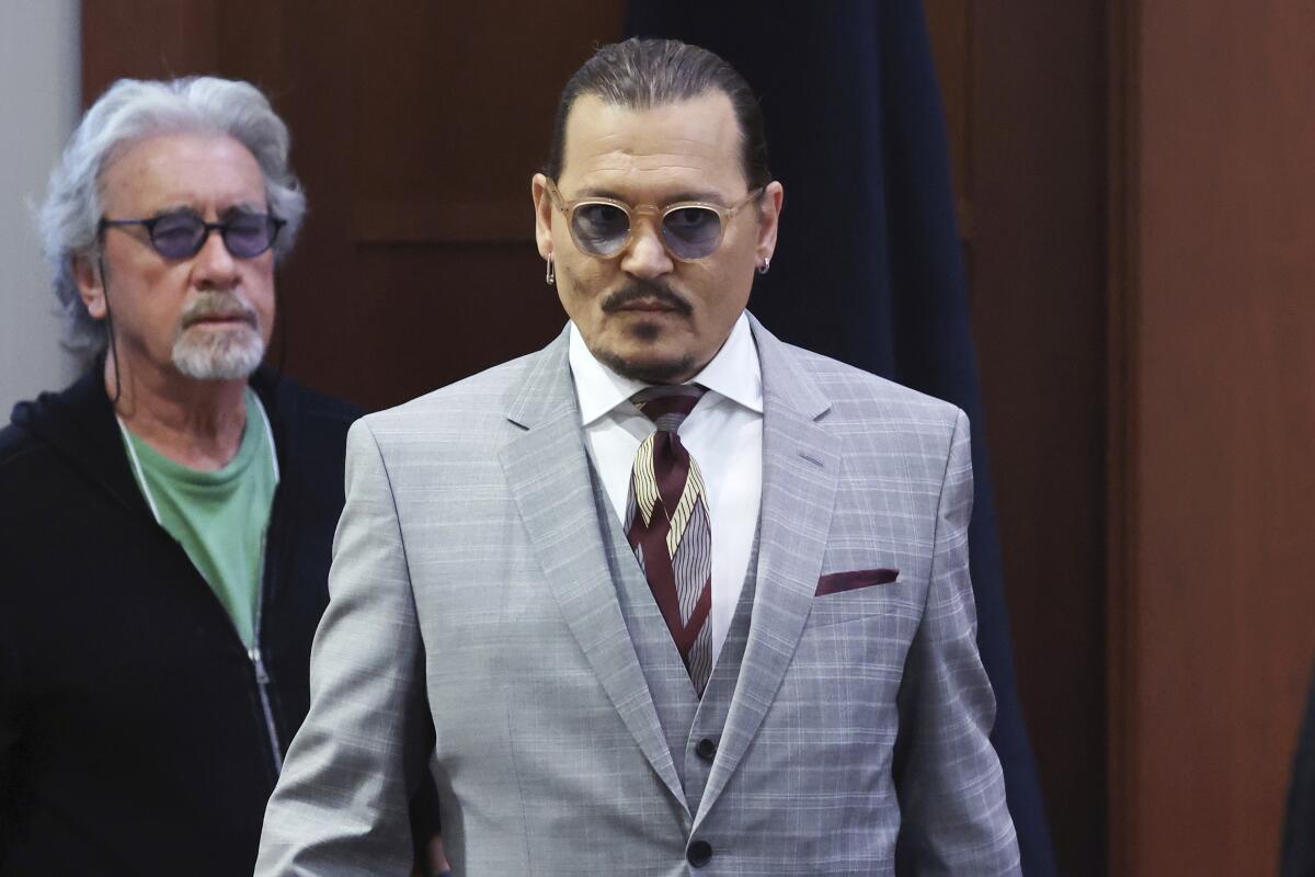 A man in a gray suit and tinted glasses walks into a courtroom with another man following him.